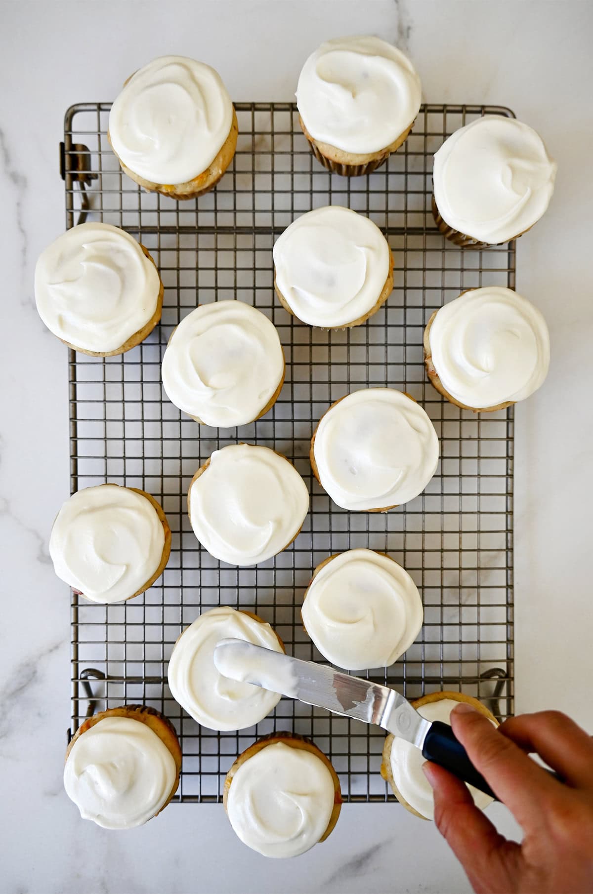 Spreading cream cheese frosting atop peach cupcakes with an offset spatula.
