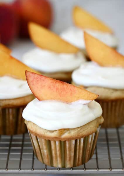 A fresh peach cupcake with cream cheese frosting, topped with a slice of peach as a garnish on a wire cooling rack.