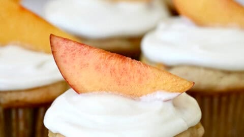A fresh peach cupcake with cream cheese frosting, topped with a slice of peach as a garnish on a wire cooling rack.