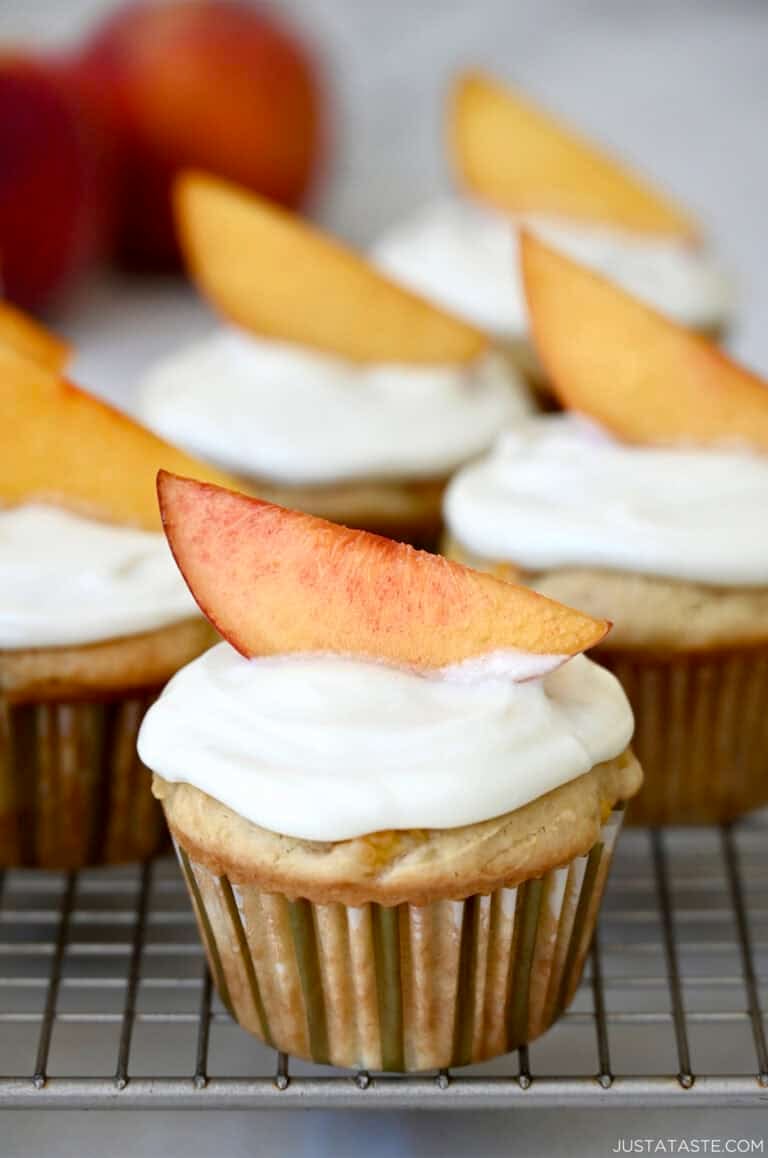 A fresh peach cupcake with cream cheese frosting, topped with a slice of peach as a garnish on a wire cooling rack.