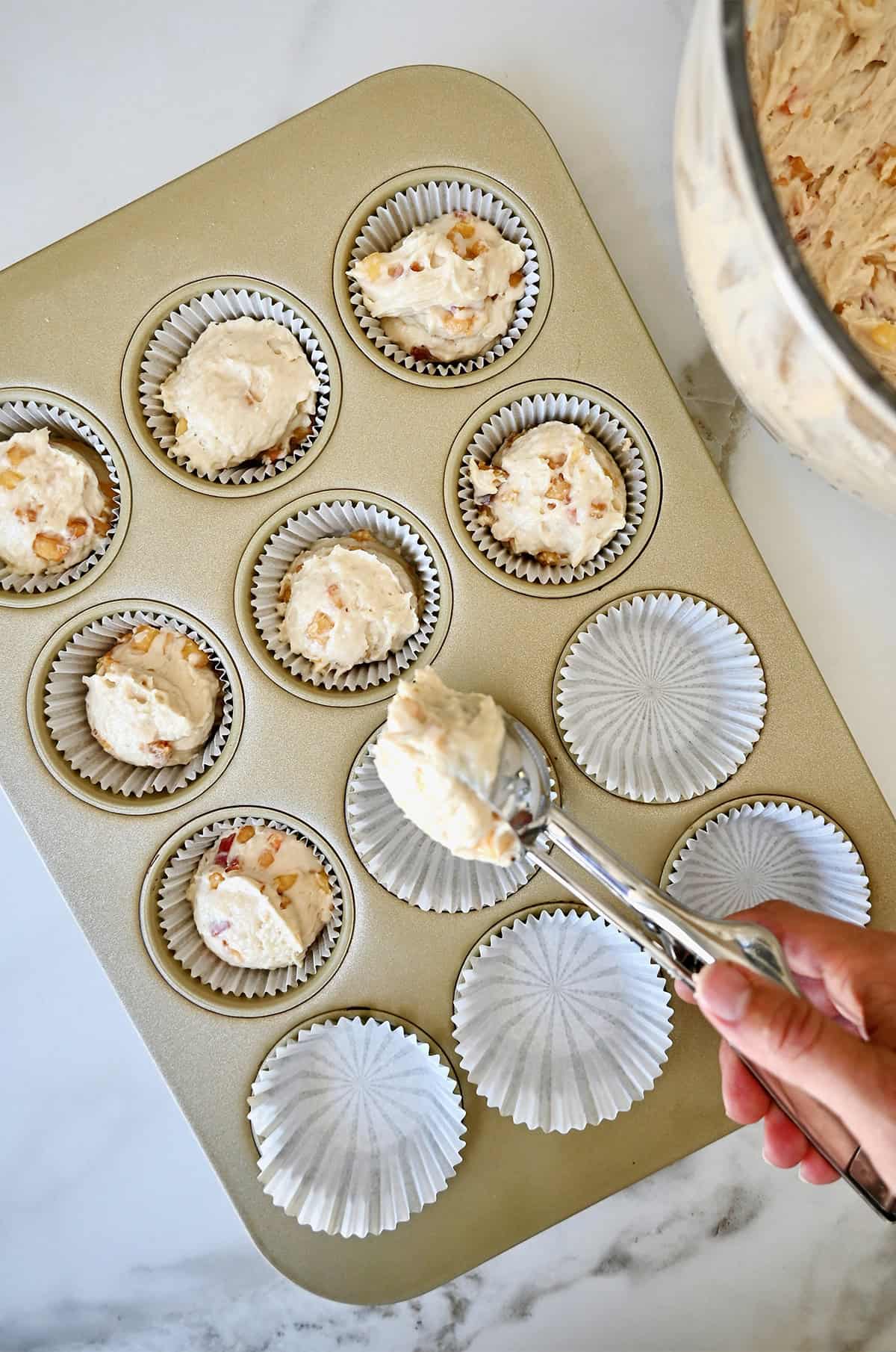 Scooping cupcake batter with diced peaches into a muffin pan with liners.