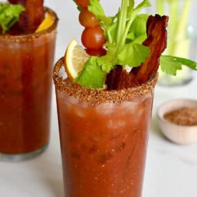 Two tall glasses filled with homemade Bloody Mary mix and garnished with crispy bacon, cherry tomatoes on a skewer, celery sticks and a lemon wedge.