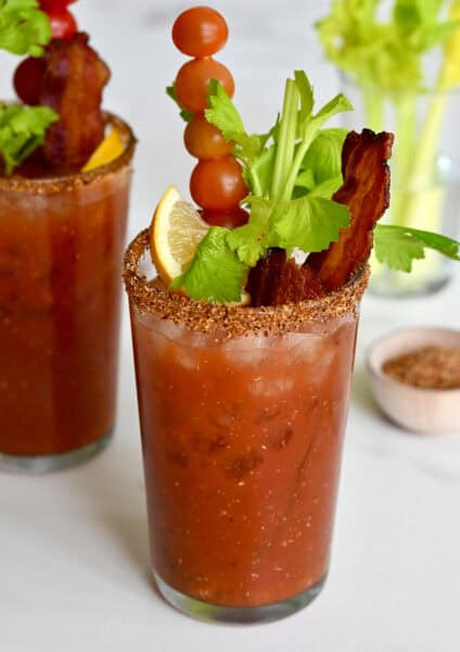 Two tall glasses filled with homemade Bloody Mary mix and garnished with crispy bacon, cherry tomatoes on a skewer, celery sticks and a lemon wedge.
