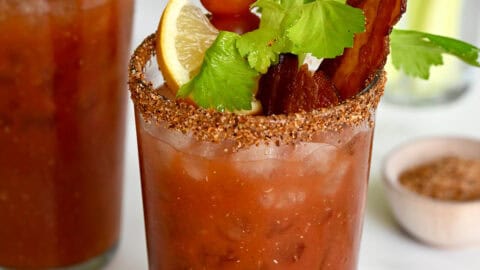 Two tall glasses filled with homemade Bloody Mary mix and garnished with crispy bacon, cherry tomatoes on a skewer, celery sticks and a lemon wedge.