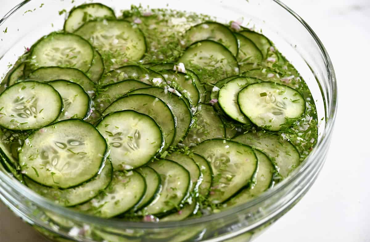 Sliced cucumbers soaking in a sugar and vinegar solution in a clear bowl.