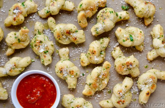 Easy Homemade Garlic Knots sprinkled with Parmesan cheese and parsley with small white bowl in bottom left corner filled with marinara sauce.