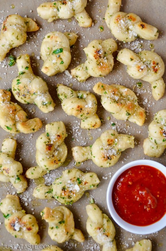 Easy Homemade Garlic Knots sprinkled with Parmesan cheese and parsley with small white bowl in bottom right corner filled with marinara sauce.