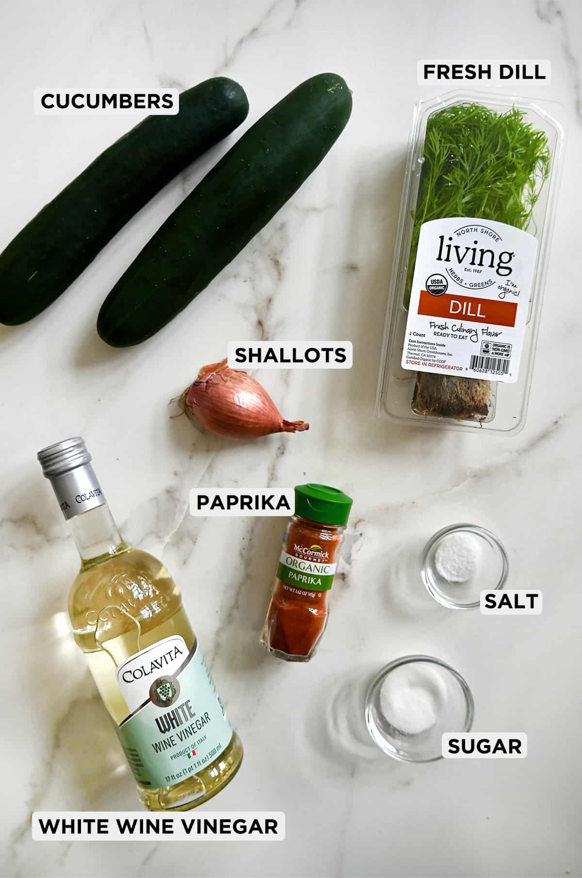 A labeled photo of ingredients for cucumber salad, including white wine vinegar, cucumbers, shallots, fresh dill, paprika, salt and sugar.