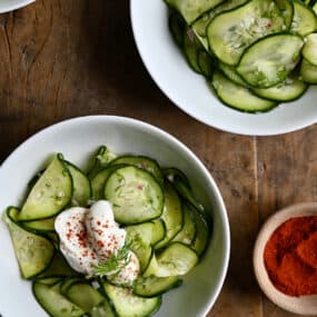 Two white bowls containing Hungarian Cucumber Salad topped with a dollop of sour cream and paprika.