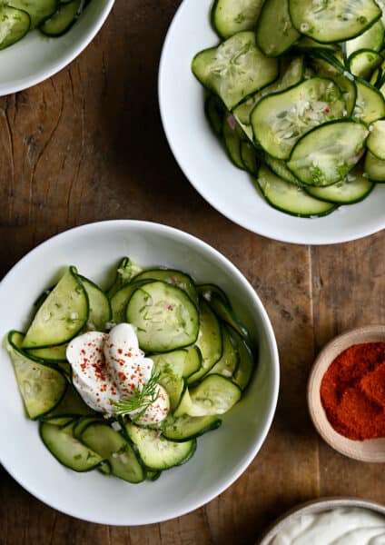 Two white bowls containing Hungarian Cucumber Salad topped with a dollop of sour cream and paprika.