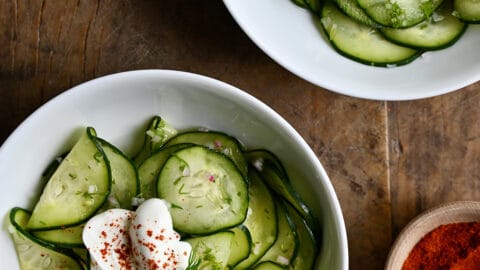 Two white bowls containing Hungarian Cucumber Salad topped with a dollop of sour cream and paprika.