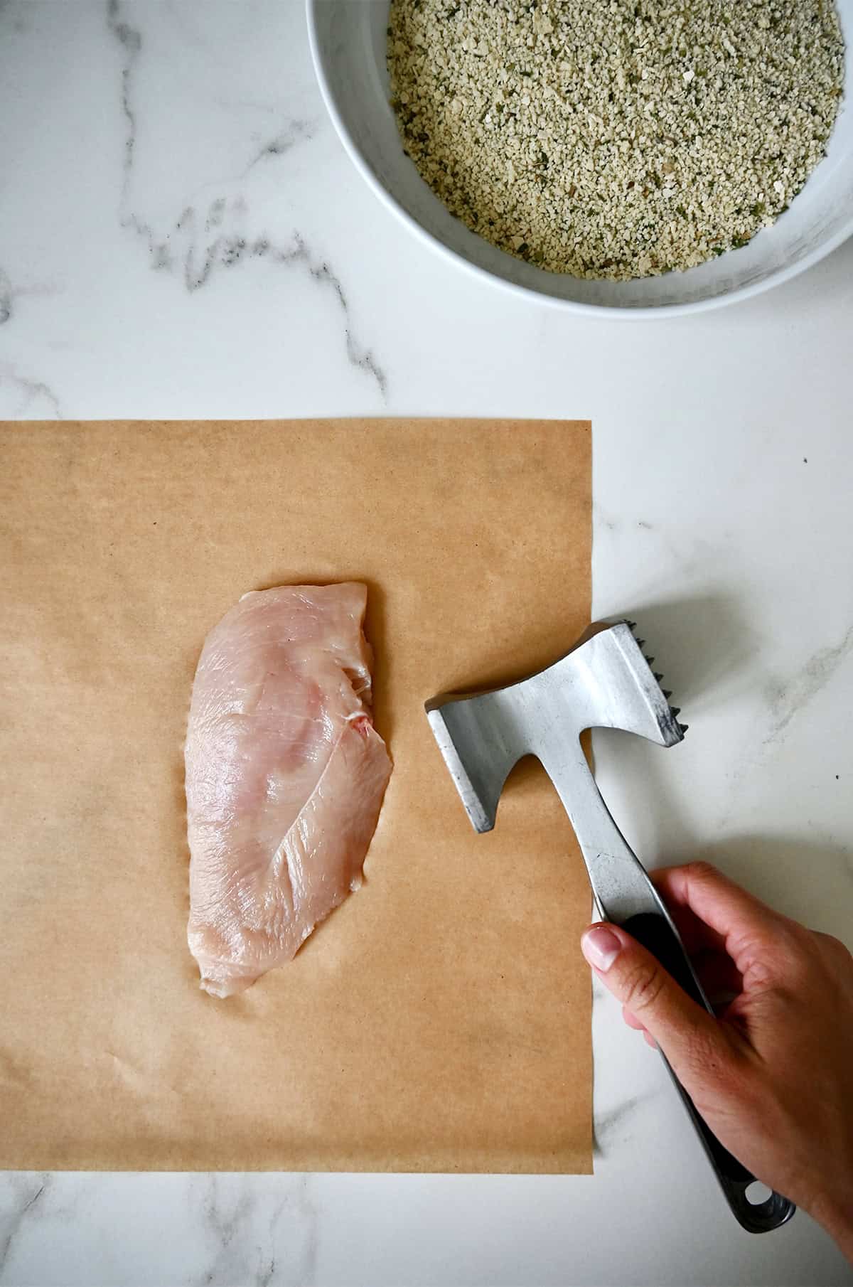 A meat mallet near a chicken breast that sits atop parchment paper.