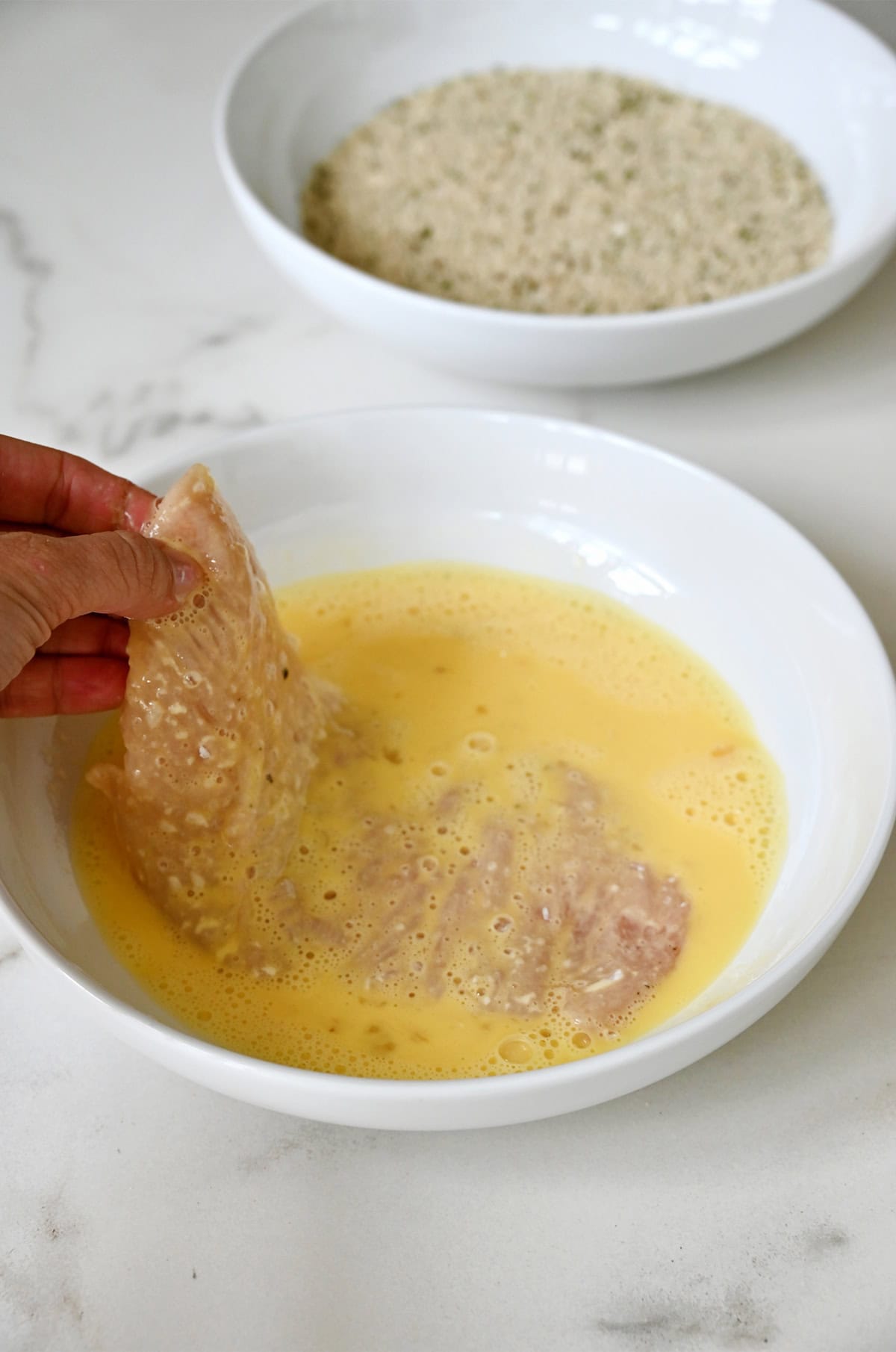 A chicken cutlet coated in flour being dipped into whisked eggs.