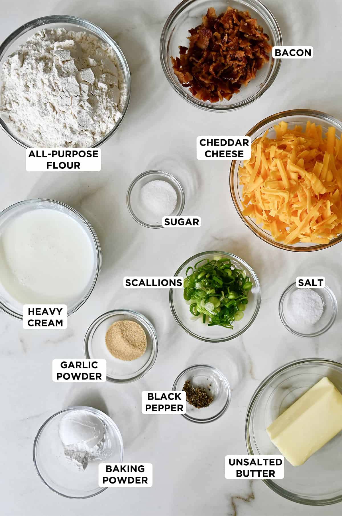 Various sizes of glass bowls containing flour, crispy bacon crumbles, shredded cheddar cheese, sugar, sliced scallions, salt, heavy cream, black pepper, butter and bacon powder.