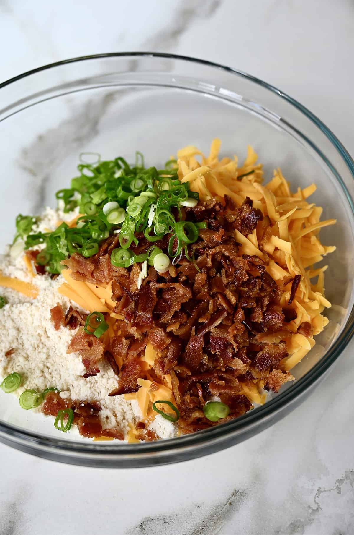 A glass bowl containing flour, crispy bacon pieces, shredded cheddar cheese and sliced scallions.