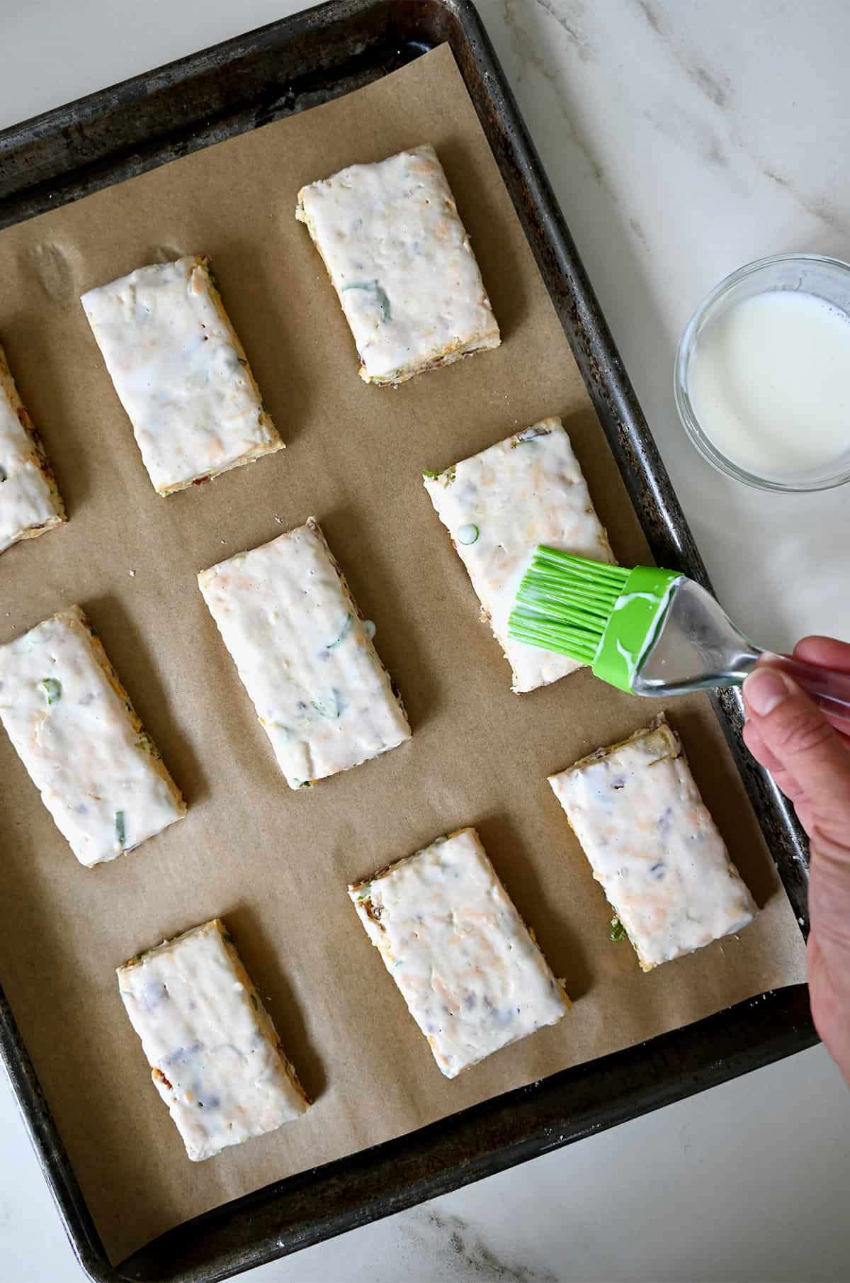 A hand holding a pastry brush applies heavy cream atop biscuits on a parchment paper-lined baking sheet.