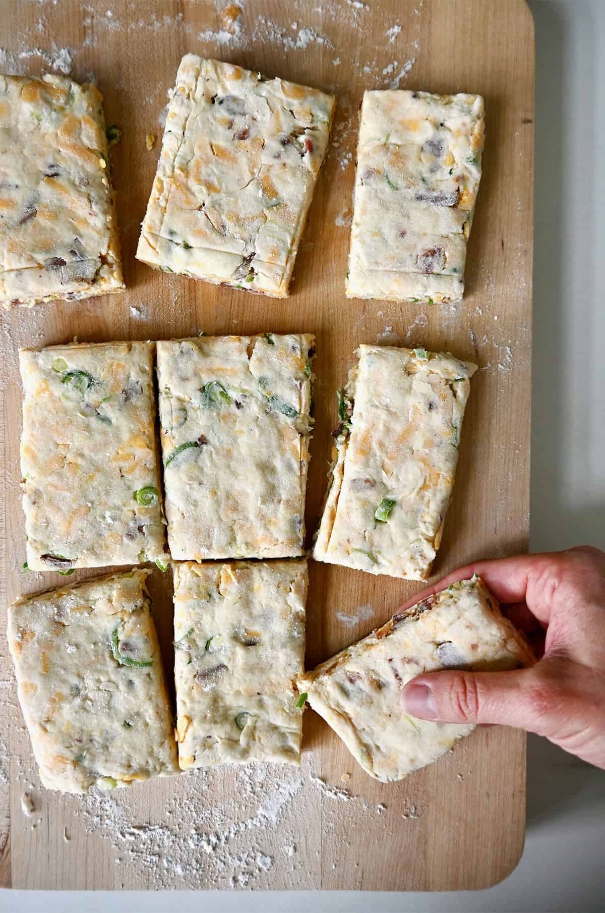 Biscuit dough cut into nine pieces on a wood cutting board with a hand holding onto one.