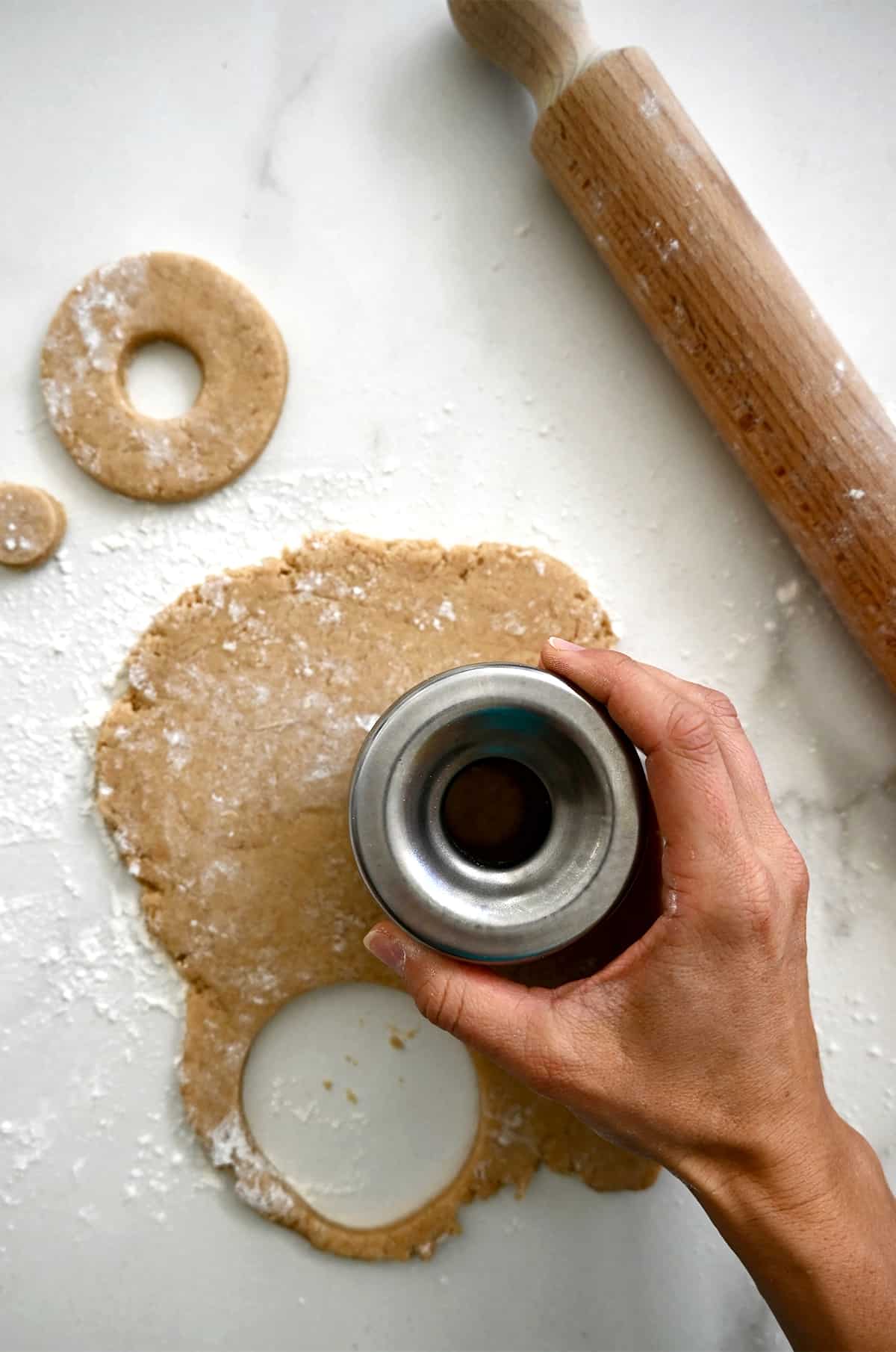 A hand holds a round cookie cutter atop doughnut dough to cut out pieces of dough to fry.