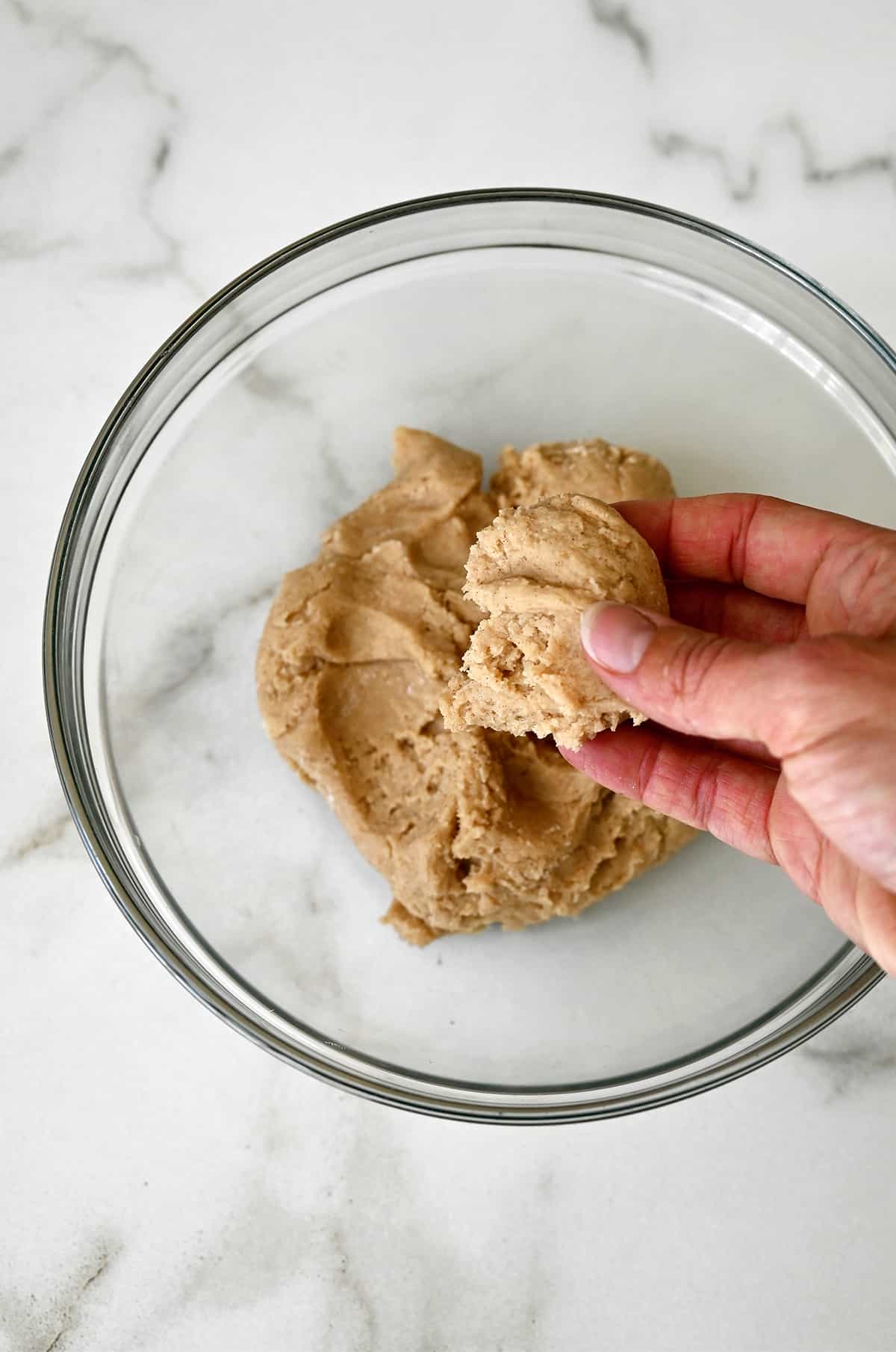 A hand holds a clump of doughnut dough over a bowl containing dough.