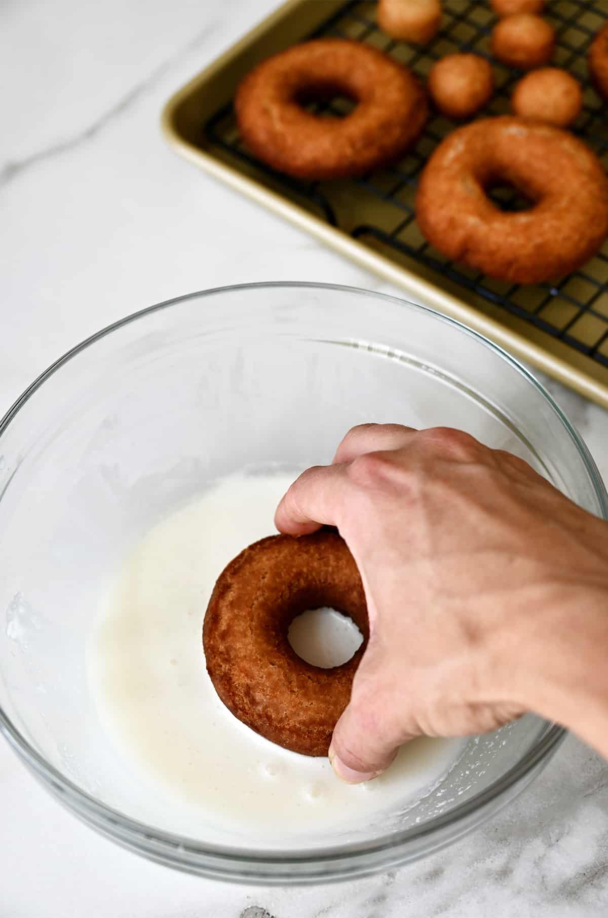A hand holding a doughnut dips it into bowl with a homemade vanilla glaze. 