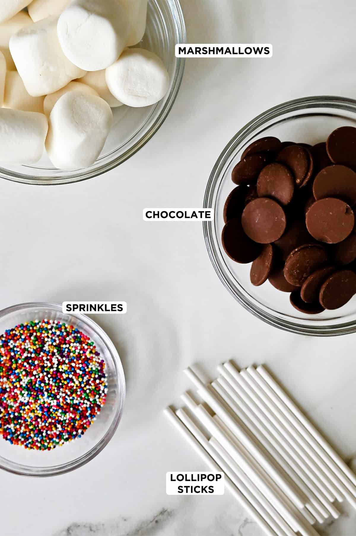 Lollipop sticks next to three bowls containing rainbow sprinkles, chocolate and marshmallows.