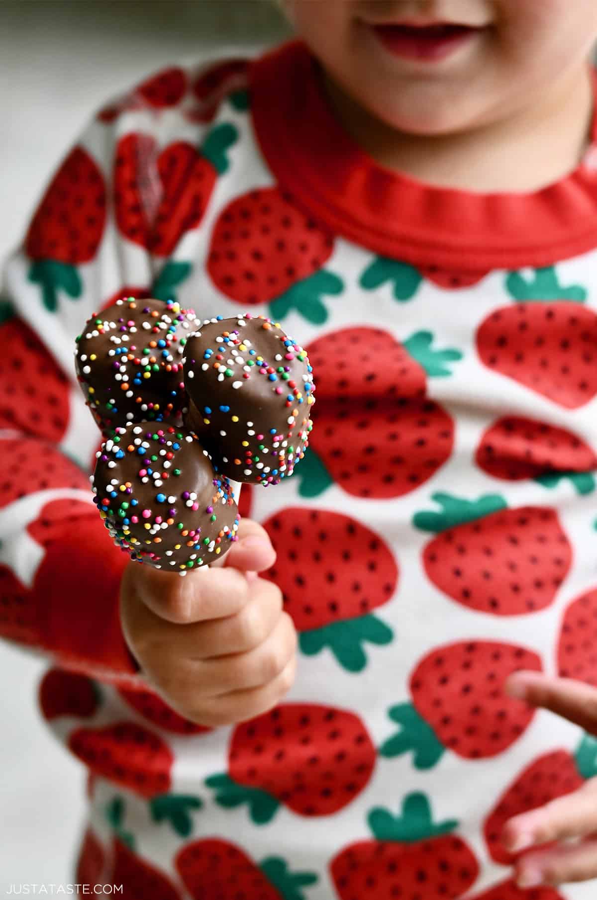A child wearing strawberry pajamas holds three chocolate marshmallow pops in her hand.