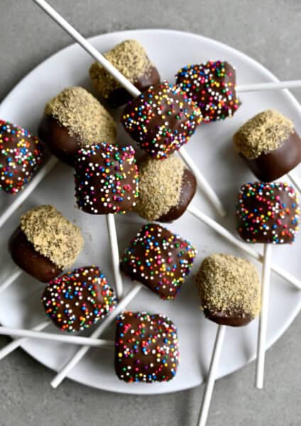 Chocolate-covered marshmallows on a stick on a serving plate. Half of the chocolate dipped marshmallows have rainbow sprinkles and half are coated in crushed nuts.