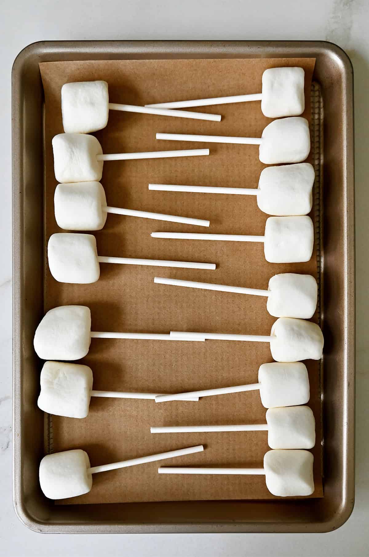 Marshmallows skewered with lollipop sticks on a parchment paper-lined baking sheet.