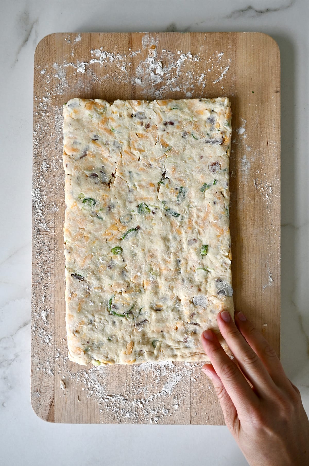 Biscuit dough with cheese, scallions and bacon shaped into a rectangle on a wood cutting board.
