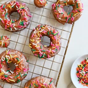 Sour cream doughnuts coated in a vanilla glaze and topped with rainbow sprinkles on a wire cooling rack.