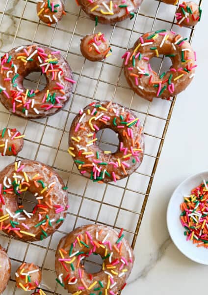 Sour cream doughnuts coated in a vanilla glaze and topped with rainbow sprinkles on a wire cooling rack.