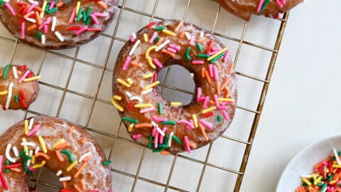 Sour cream doughnuts coated in a vanilla glaze and topped with rainbow sprinkles on a wire cooling rack.