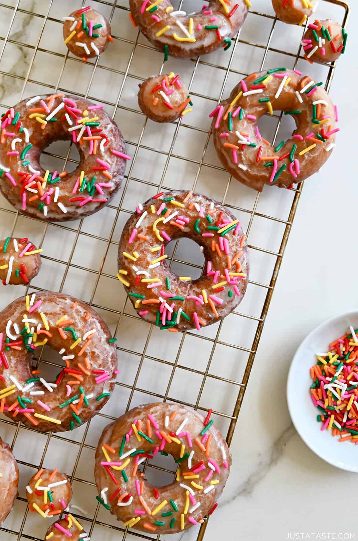 Sour cream doughnuts coated in a vanilla glaze and topped with rainbow sprinkles on a wire cooling rack.