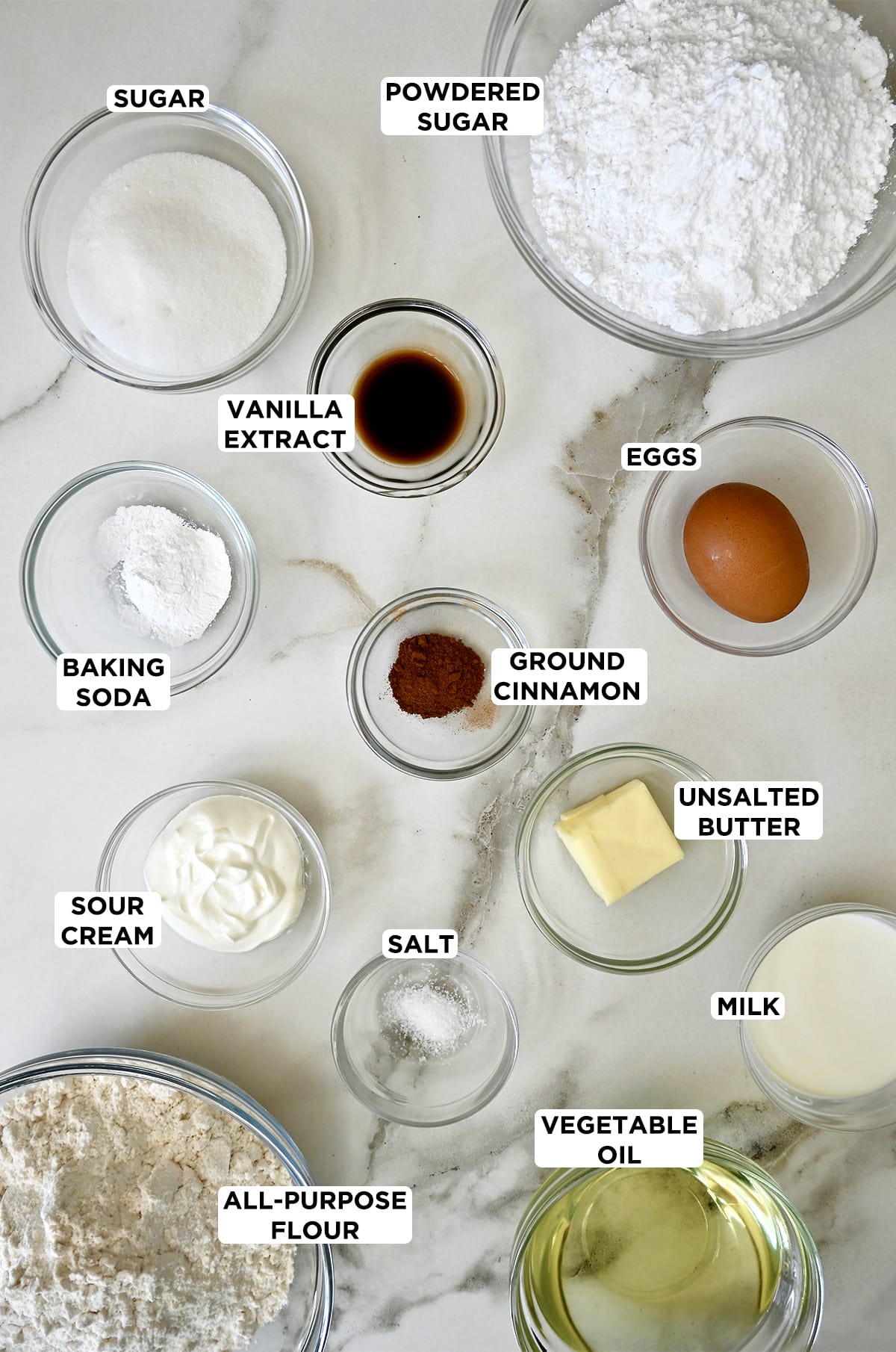 Various sizes of clear bowl containing powdered sugar, an egg, unsalted butter, milk, vegetable oil, salt, flour, sour cream, baking soda, vanilla extract and granulated sugar.