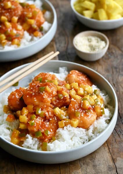 Pineapple shrimp with chopped green peppers tossed in a sweet and spicy sauce atop coconut rice in a bowl with chopsticks.
