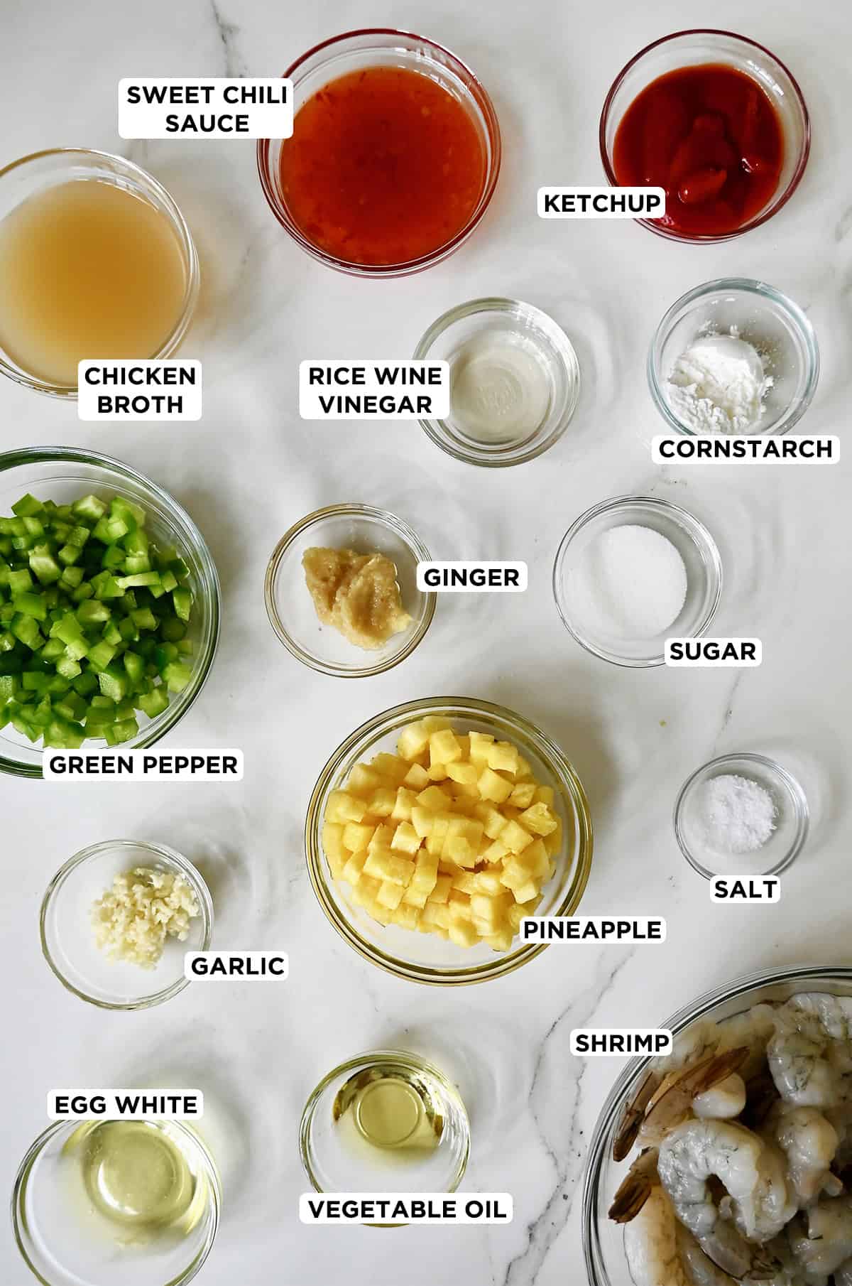 Various sizes of glass bowls containing the ingredients needed to make pineapple shrimp: sweet chili sauce, ketchup, cornstarch, rice wine vinegar, sugar, minced ginger and garlic, salt, chopped pineapple and green peppers, shrimp, chicken broth, and vegetable oil for frying.