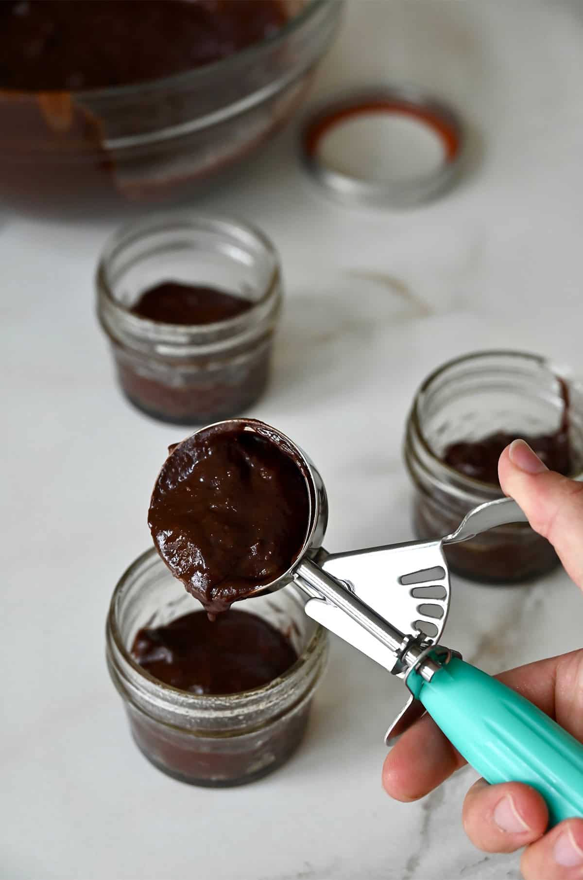 An ice cream scoop with brownie batter over small mason jars.
