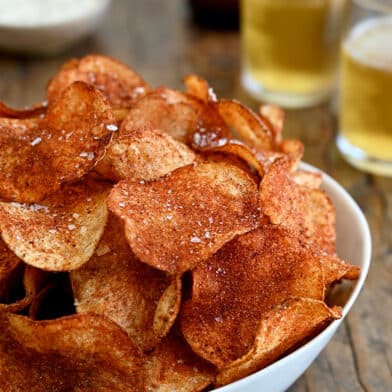 A white serving bowl containing Homemade Barbecue Potato Chips with two glasses filled with beer in the background