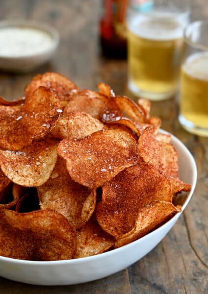 A white serving bowl containing Homemade Barbecue Potato Chips with two glasses filled with beer in the background