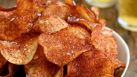 A white serving bowl containing Homemade Barbecue Potato Chips with two glasses filled with beer in the background