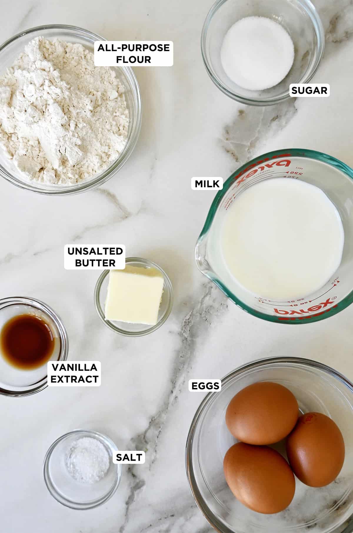 Glass bowls containing ingredients to make a Dutch baby pancake, including flour, sugar, milk, eggs, butter, salt and vanilla extract.