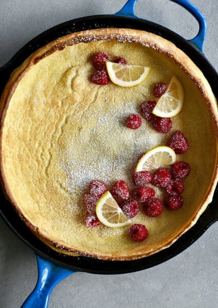 Dutch baby pancake in a cast iron skillet topped with lemon slices, fresh raspberries and a dusting of powdered sugar.