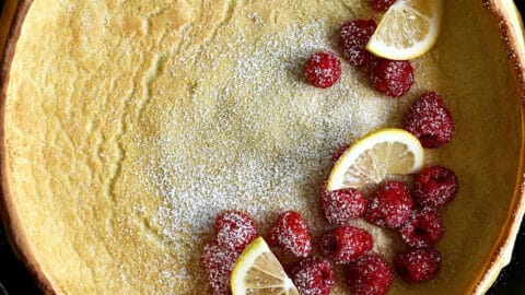 Dutch baby pancake in a cast iron skillet topped with lemon slices, fresh raspberries and a dusting of powdered sugar.