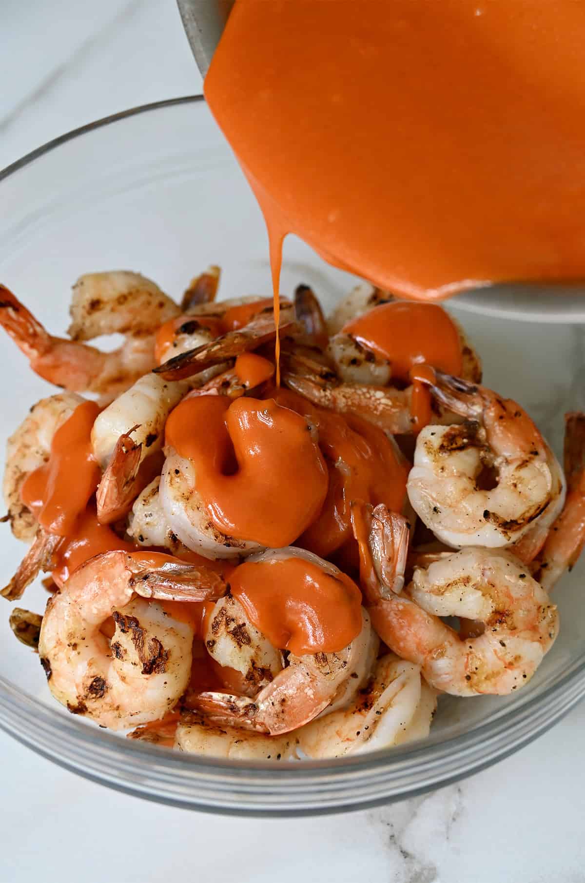 Buffalo sauce being poured over charred shrimp in a glass bowl.