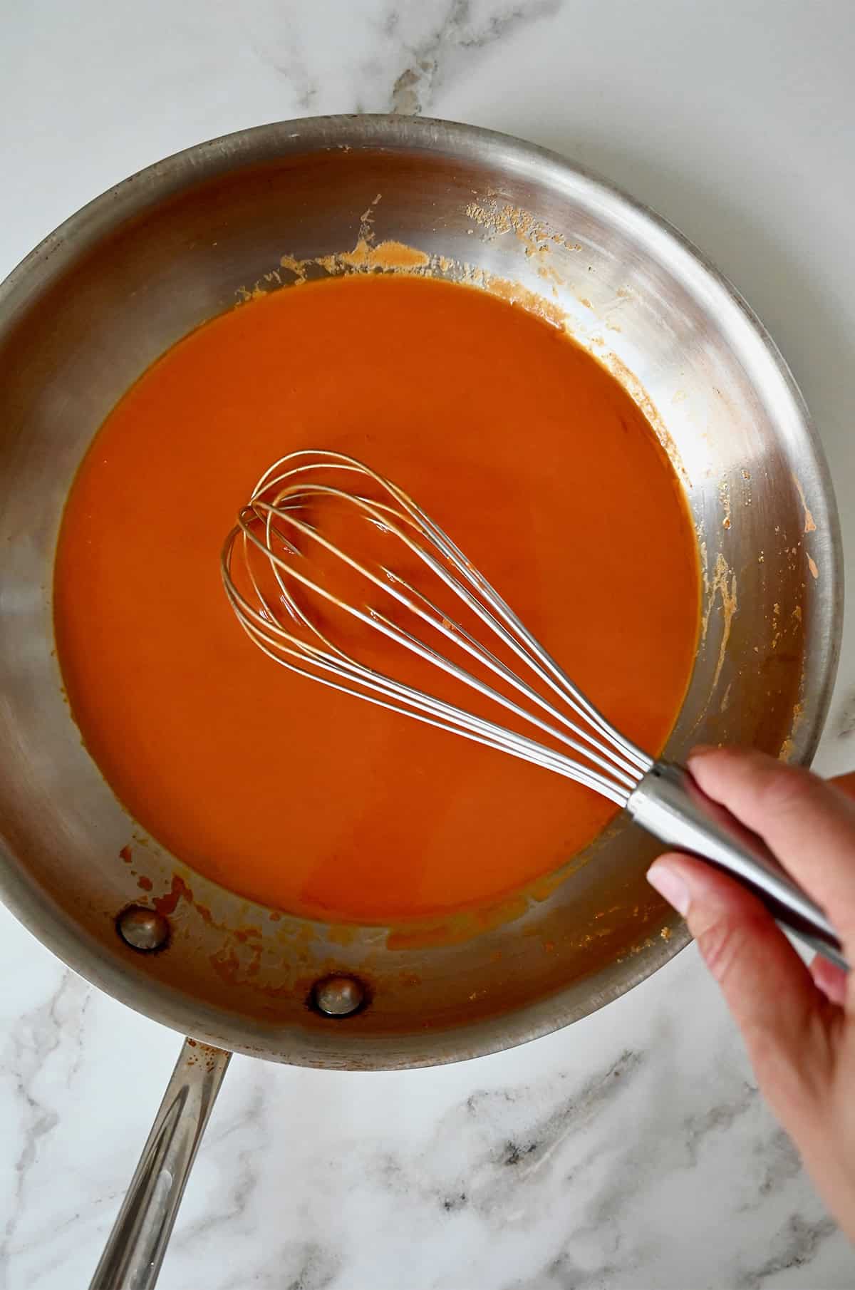 Buffalo sauce in a sauté pan being stirred with a wire whisk.