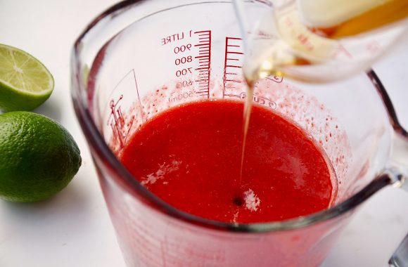 Tequila being poured into measuring cup containing strawberry purée next to limes 