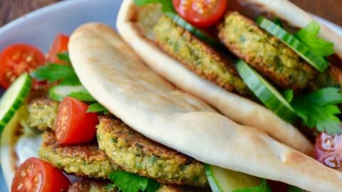 Easy Homemade Falafel tucked inside pita bread with Tahini Sauce, cucumbers, tomatoes and parsley. Small blue blue with cherry tomatoes and white plate with chopped parsley in background.