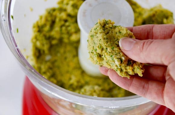 Hand holding shaped falafel with food processor containing falafel mixture in background.
