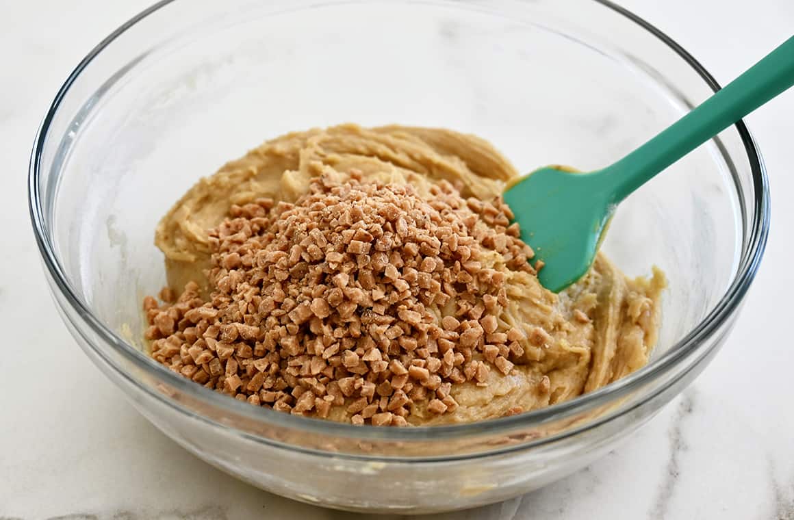 A teal spatula in a clear bowl that contains blondie batter and toffee bits.