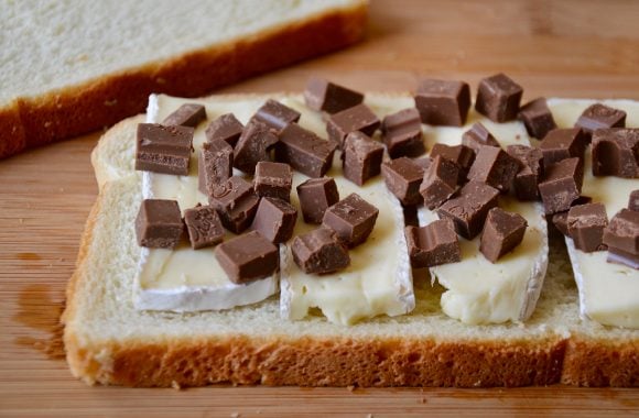 A slice of white bread topped with brie cheese and chopped chocolate on a cutting board